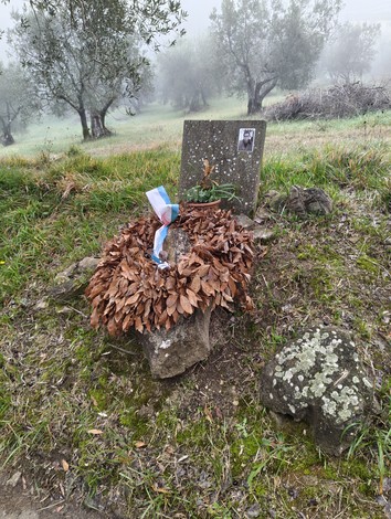 Cippo commemorativo avvolto da foglie secche, situato in un paesaggio verdeggiante e nebbioso.