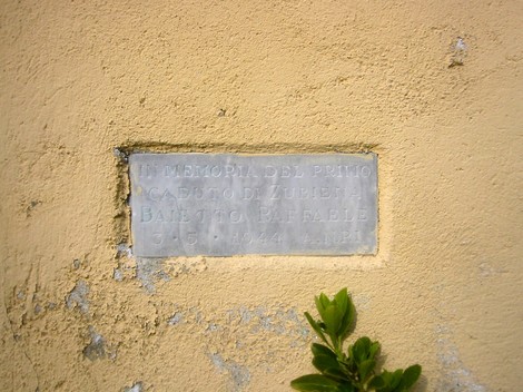 Lapide commemorativa dedicata a Baietto, situata su un muro giallo. Stile semplice e leggermente danneggiata.