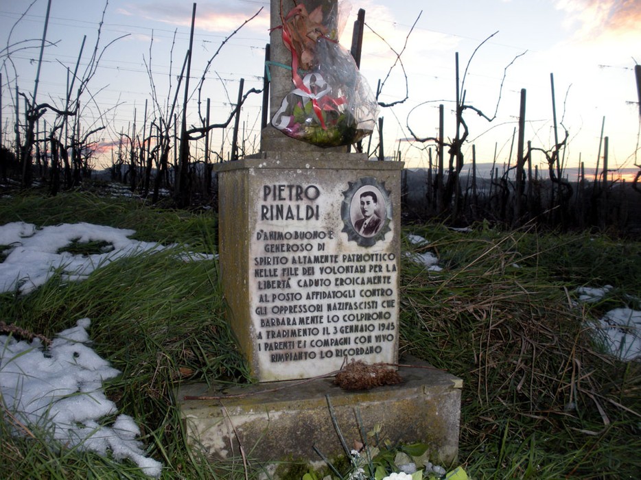 Cippo commemorativo dedicato a Rinaldi, in un paesaggio di vigne e neve al tramonto.