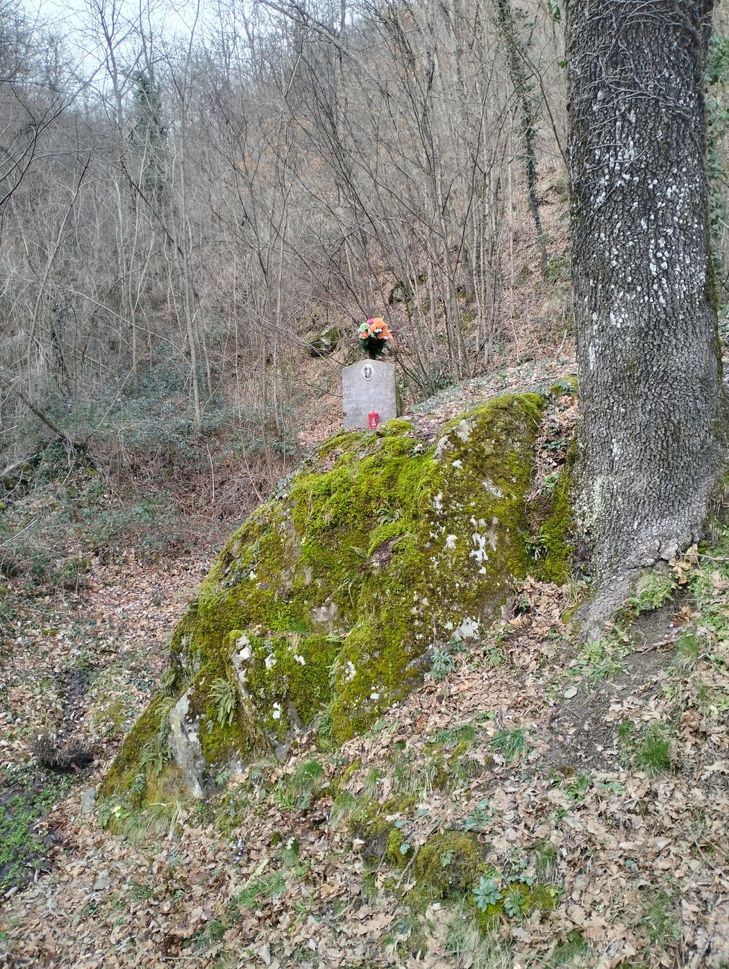 Cippo commemorativo adornato con fiori su una roccia, circondato da alberi e vegetazione in un'ambientazione naturale.