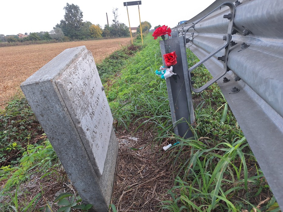 Cippo in pietra con iscrizione sul ciglio di una strada, accanto a un guardrail con fiori rossi.