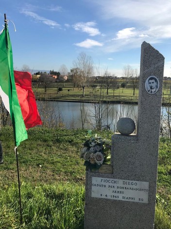 Cippo commemorativo dedicato a Diego Fiocchi, caduto per bombardamento aereo. Paesaggio sereno con bandiera italiana.