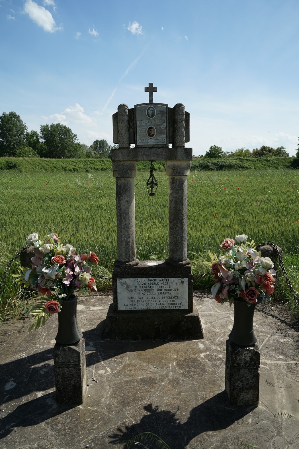 Cippo commemorativo con fiori in un campo verde sotto il cielo blu. Un simbolo di memoria e rispetto.