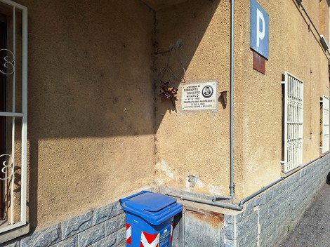 Lapide commemorativa a Ferrante, affissa su un muro di edificio con fondo colorato e contenitore blu.