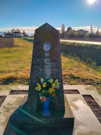 Cippo commemorativo con fiori, situato lungo una strada panoramica, immerso nella natura.