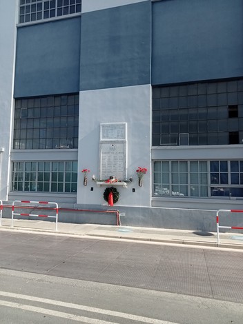 Lapide con iscrizione sulla facciata di un edificio industriale, ornata da fiori rossi e una corona.