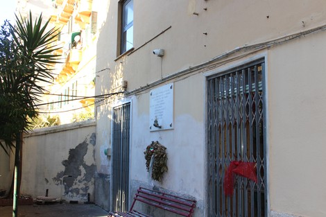 Lapide commemorativa affissa a un muro, decorata con una corona floreale, in una zona urbana.