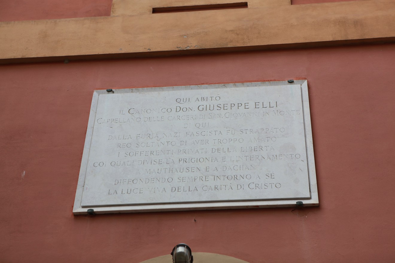 Lapide commemorativa dedicata a Don Giuseppe Elli, situata su una parete in un contesto architettonico.