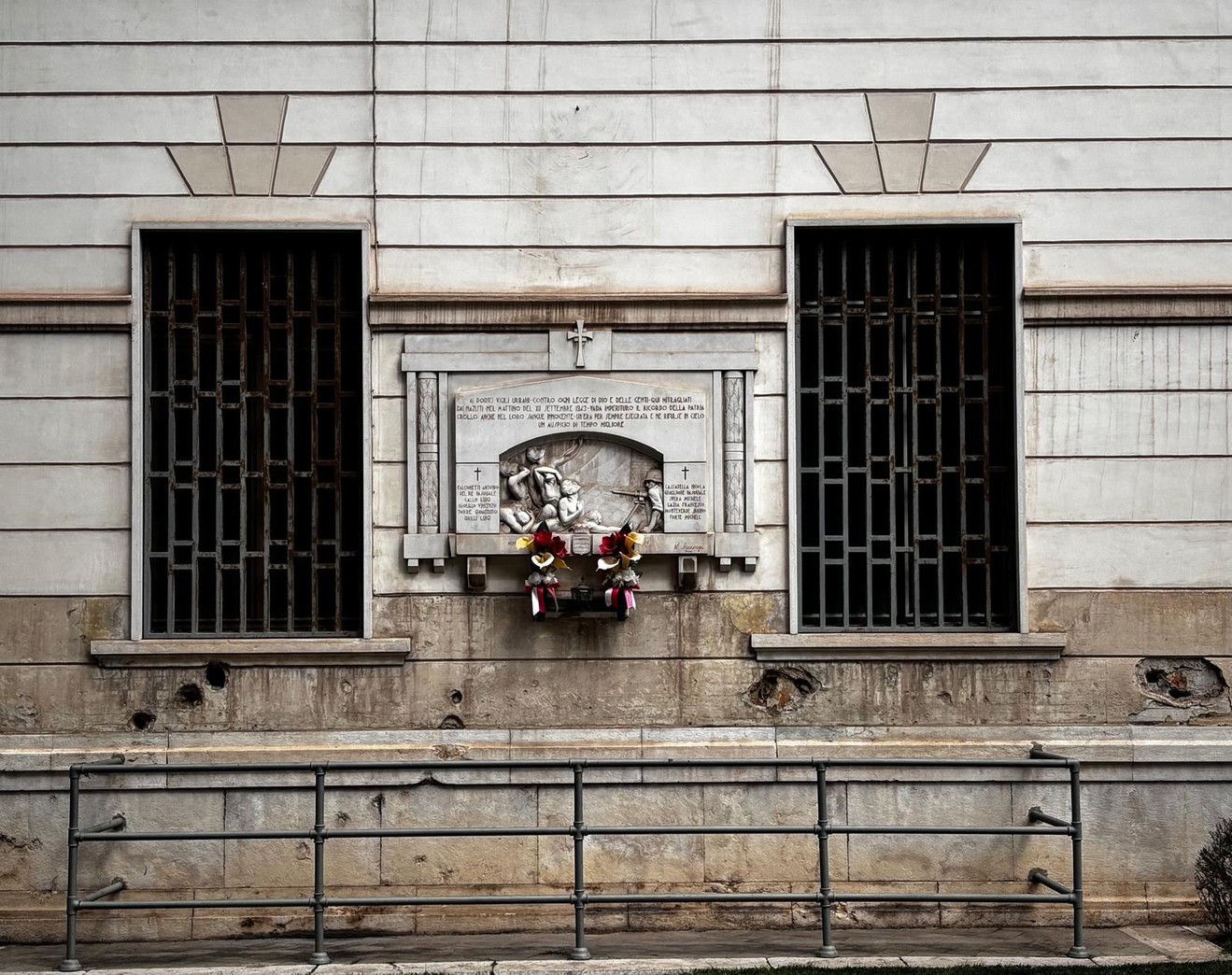 Lapide commemorativa sull'ex palazzo delle poste, con struttura architettonica sobria e fiori deposti.