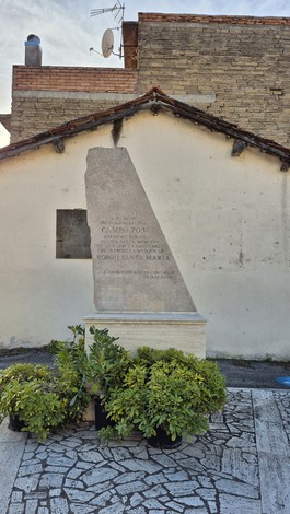 Monumento al Campo PG54, con una struttura in pietra e vegetazione circostante, su sfondo di un edificio.