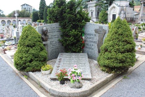 Tomba commemorativa con monumento ai caduti, circondata da piante e fiori, in un cimitero.