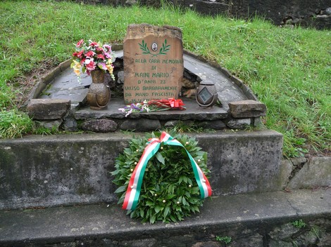Cippo commemorativo dedicato a Maini Mario, decorato con fiori e un cesto di alloro, immerso nel verde.
