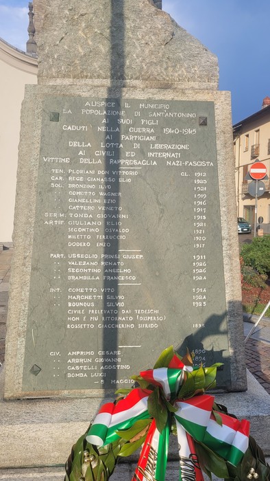 Lapide in pietra con lunga iscrizione, posta su un basamento in una piazza, con corona tricolore ai piedi.