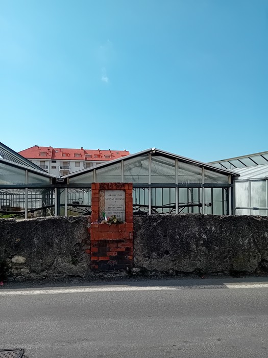 Lapide in pietra visibile accanto a serra, con edifici residenziali sullo sfondo e cielo sereno.