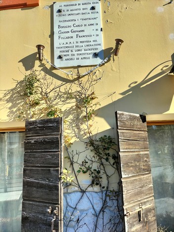 Lapide commemorativa a Bonaldo, Gardin e Follador su una parete con porte in legno e vegetazione circostante.