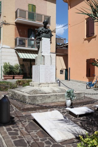 Monumento ai Caduti con statua e lastre commemorative in un'area urbana, circondato da edifici e vegetazione.