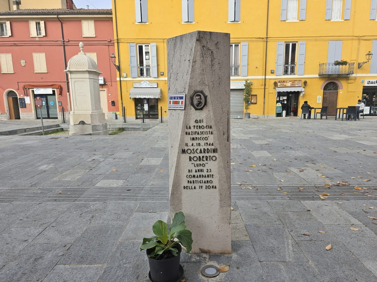Cippo commemorativo dedicato a Moscardini, situato in una piazza con edifici colorati sullo sfondo.