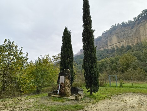 Monumento agli uccisi di Rio Conco, immerso nella natura con cipressi e scogliere sullo sfondo.