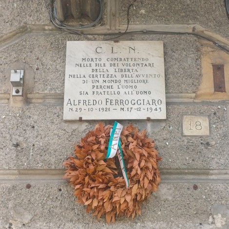 Lapide in marmo su parete esterna, con iscrizione dedicatoria e corona di foglie appesa sotto.