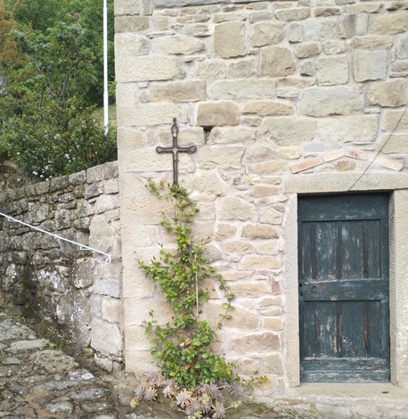 Croce in ferro battuto affissa a un muro di pietra, con una pianta rampicante che cresce vicino a una porta in legno.