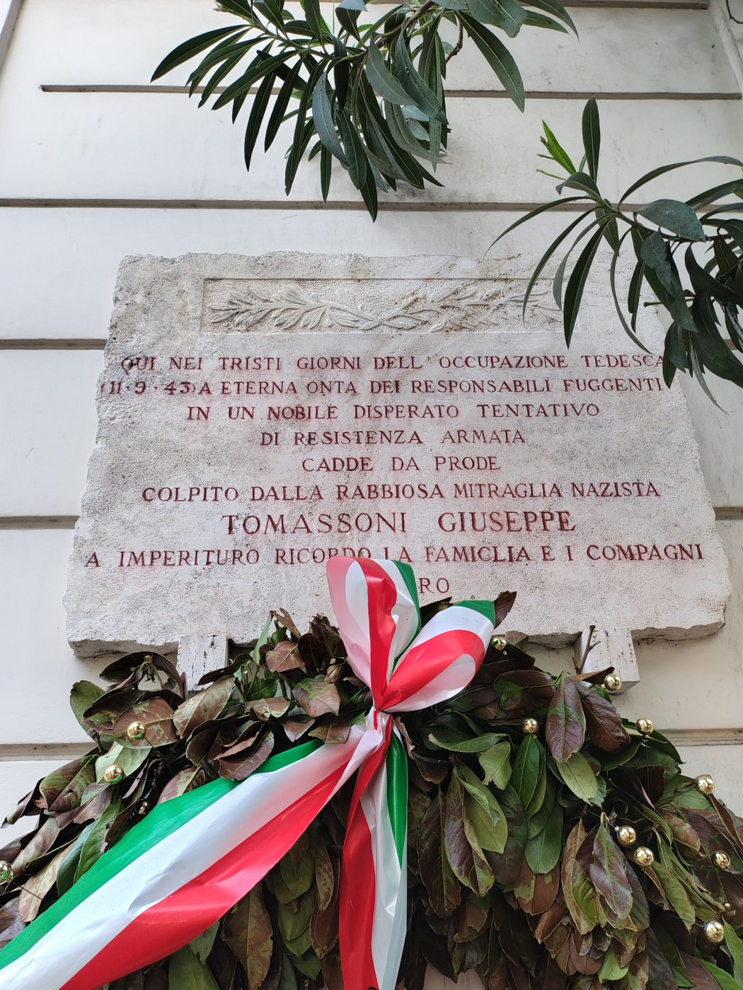 Lapide commemorativa dedicata a Giuseppe Tomassoni, con corone di fiori e nastri tricolore.