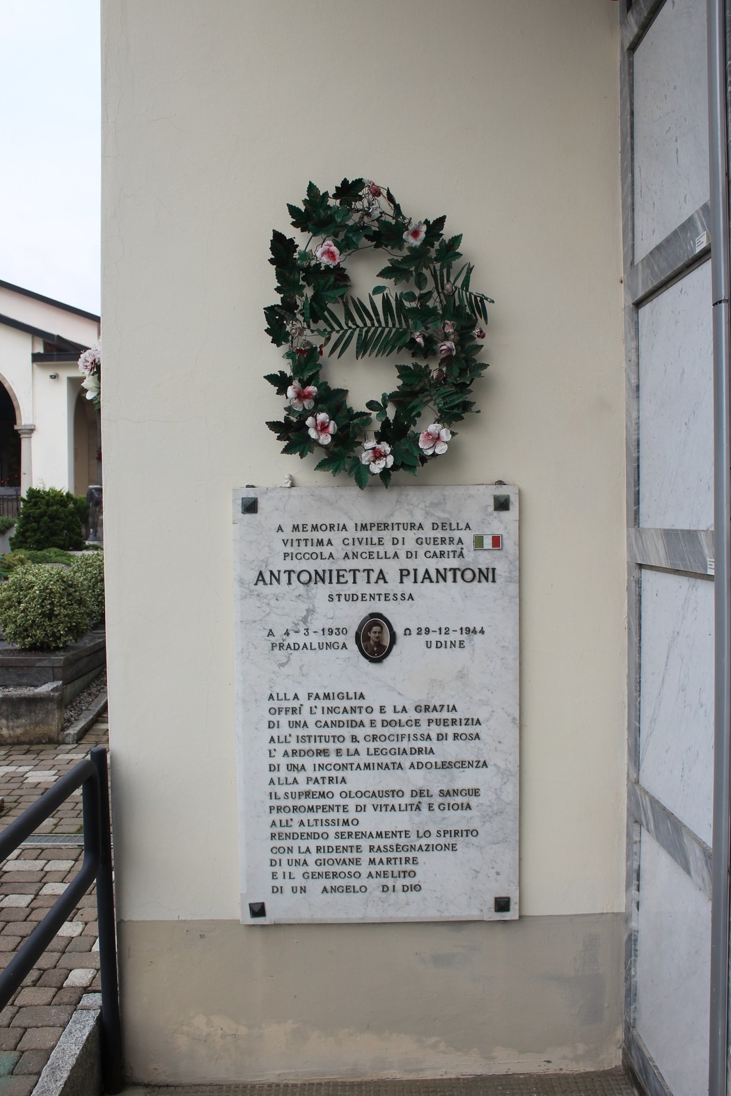 Lapide commemorativa in memoria di Antonietta Piantoni, adornata da una corona di fiori.