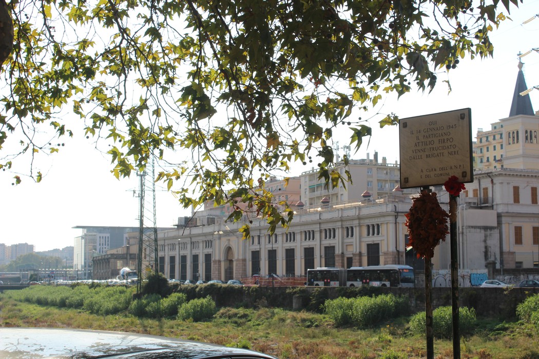 Lapide commemorativa in un contesto urbano, con edifici e vegetazione circostante visibili sullo sfondo.