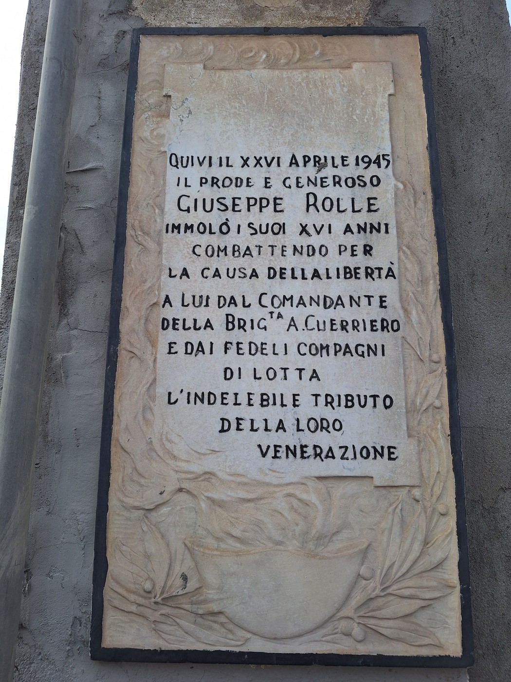 Lapide in marmo su parete con cornice decorata e iscrizione, in basso rilievo di alloro e scudo.