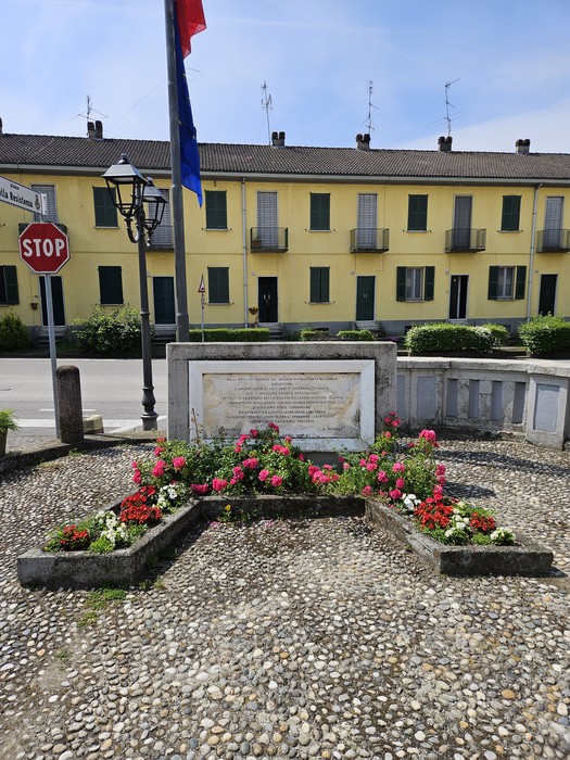 Monumento alla Brigata Balladore, circondato da fiori e cartelli stradali, in un contesto urbano.